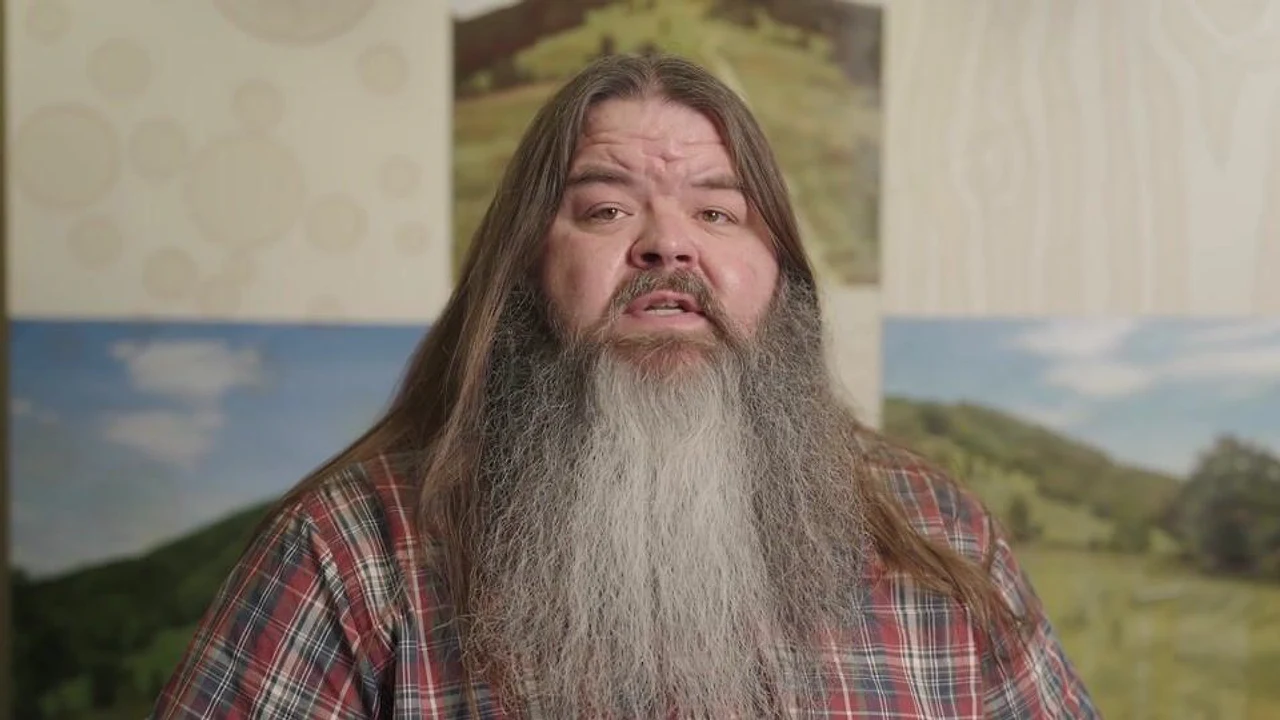 A long-haired, bearded man speaks on camera in front of landscape prints