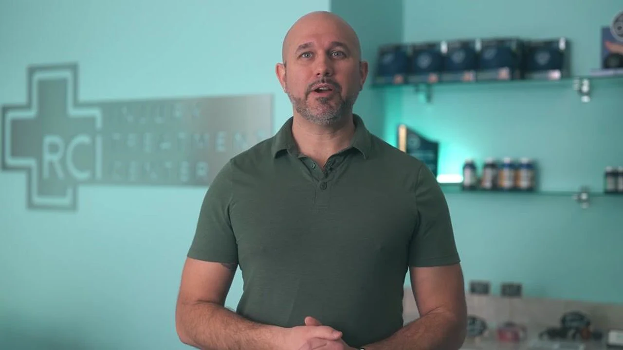 A man speaks on camera inside a teal-lit chiropractic clinic