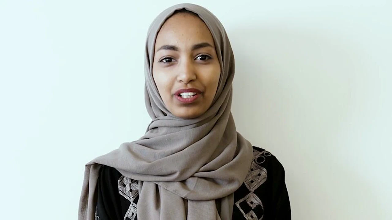 A woman in a gray headscarf speaks directly to camera against a white wall